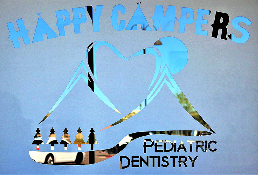 The image shows a sign with the text Happy Campers at the top, followed by a stylized graphic of a mountain peak with trees and a blue sky background. Below this graphic are two smaller images one on the left depicts a pine tree and the other on the right is a graphic of a mountain peak with clouds. The sign also includes text that reads Pediatric Dentistry at the bottom, indicating the name of the dental practice.